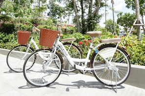 Una bicicleta blanca con cesta y asiento, estacionada sobre una superficie pavimentada con follaje al fondo.