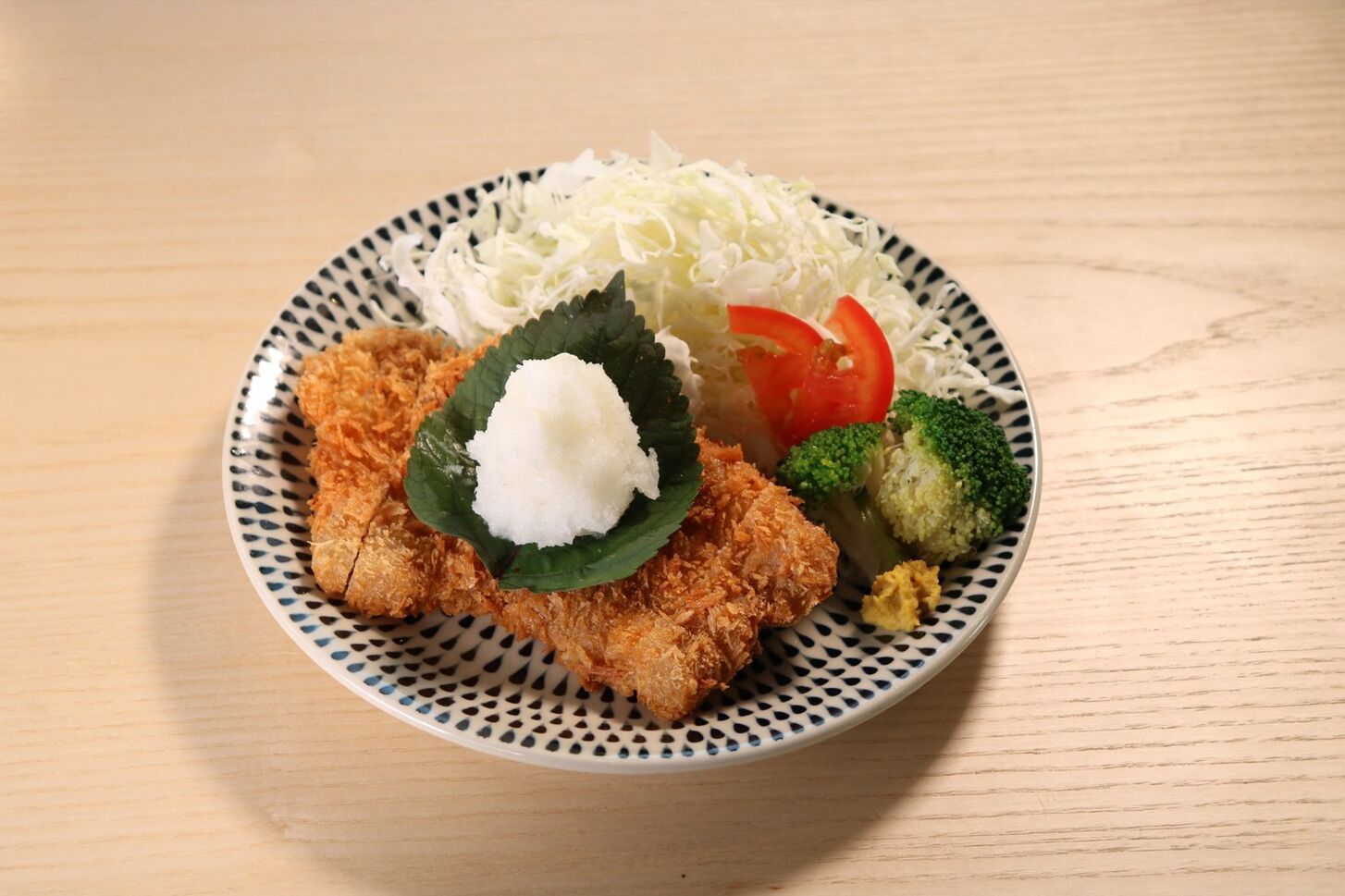 A plate with breaded cutlet, shredded cabbage, tomato slices, broccoli, and a dollop of white sauce on a patterned plate.