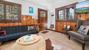 Un interior de cabaña acogedor con un sofá azul oscuro, un sillón de cuero, una mesa de centro de madera y un televisor montado en la pared.