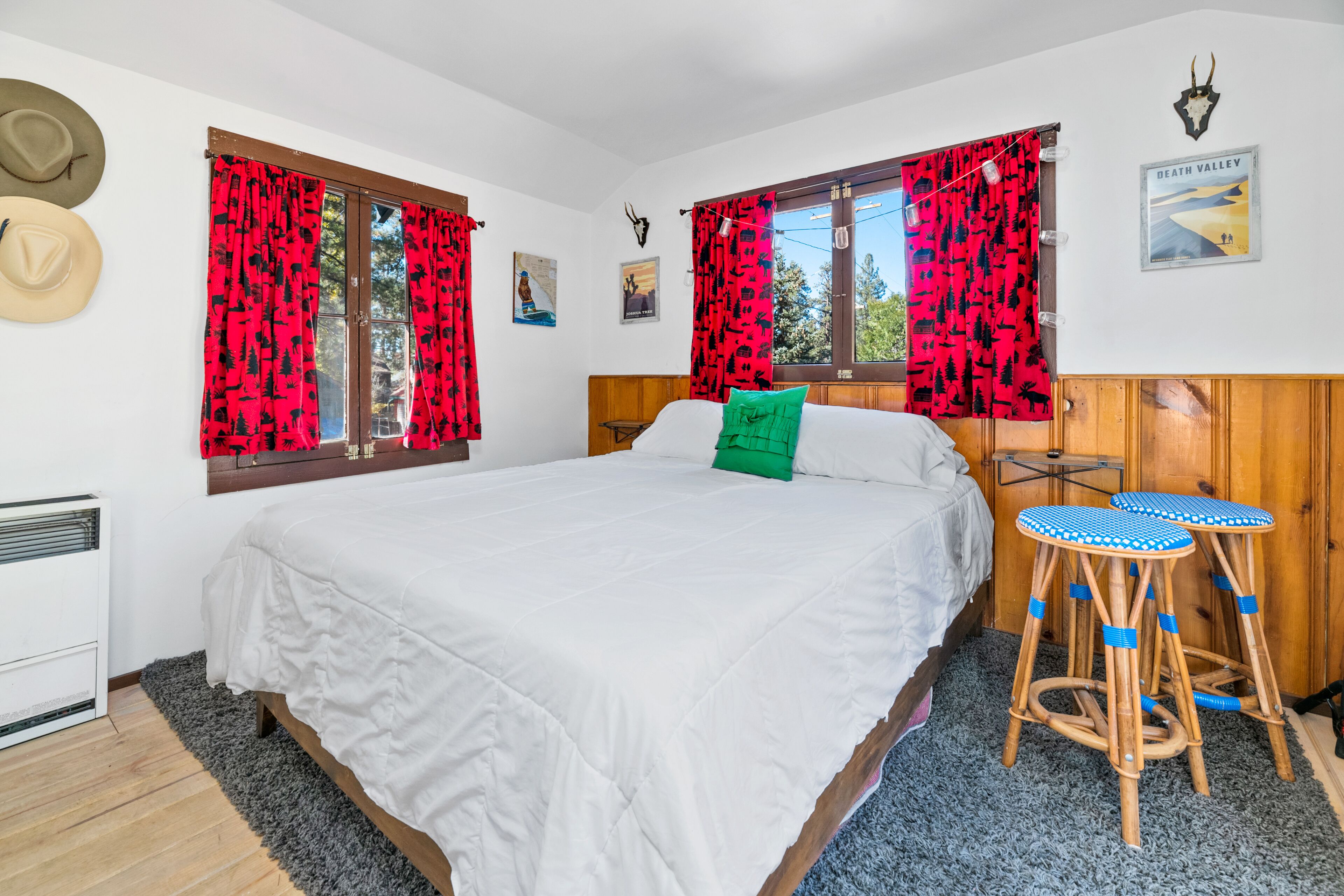 Camera da letto con un letto grande, due finestre con tende rosse a motivi decorativi, una testiera in legno, un cuscino verde e due sgabelli blu e bianchi.