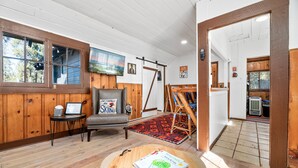 Una habitación acogedora con paredes de madera, un ventanal grande, un sillón gris, una mesita con un libro y controles remotos, y una alfombra roja y negra.