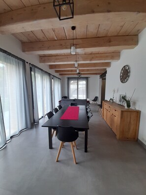 A spacious room with a long table, wooden ceiling beams, and large windows with sheer curtains.