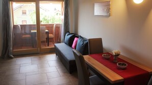 Une pièce chaleureuse avec un canapé, une table à manger agrémentée d’un chemin de table rouge, et une vue sur l’extérieur, accessible par une porte vitrée, donnant sur un espace extérieur avec une terrasse en bois et des chaises.
