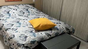 Un dormitorio con una cama, una almohada amarilla, un banco, un armario de madera y un cuadro floral en la pared.