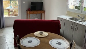 Un comedor con una mesa redonda, cuatro sillas y un mantel rojo. En el fondo, hay un televisor sobre una mesita y una ventana con un cortinaje decorativo.