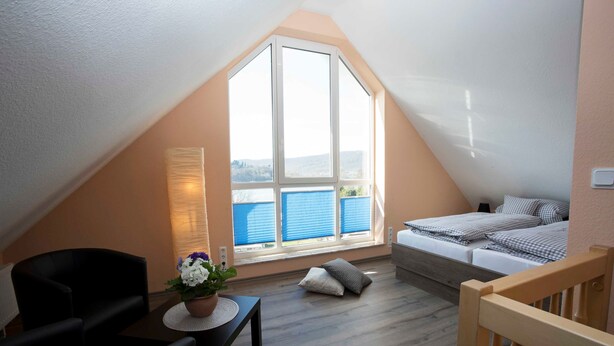 Un dormitorio acogedor en el ático con un ventanal grande, una cama, una mesita con una planta y una silla.