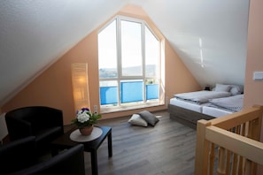 A cozy attic bedroom with a large window, a bed, a small table with a plant, and a chair.