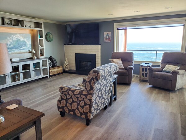 A living room with a fireplace, a sofa, armchairs, a TV, and a view of the ocean. - "By The Sea" Oceanfront Updated Rainbow Rock Condo, Spectacular Views, 1st Floor (Brookings, OR 97415)