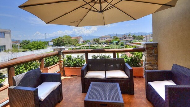 A balcony with wicker furniture and a large umbrella, overlooking a residential area with houses and greenery.