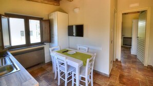 A compact kitchen with a dining table, white chairs, a TV mounted on the wall, and a window providing natural light.