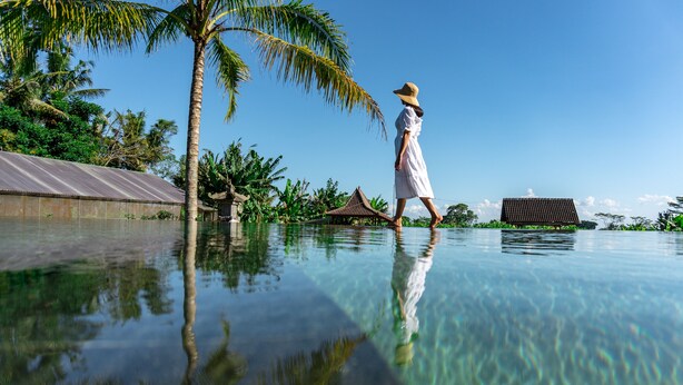 A person walking on a clear pool with a palm tree and a traditional building in the background. - Alam Kawi Ubud Resort & Spa (Ubud)