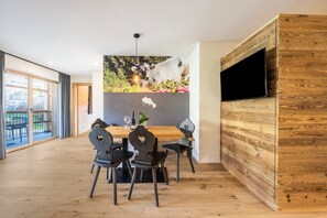 Un comedor con mesa de madera, cuatro sillas, un televisor montado en la pared y un ventanal grande.