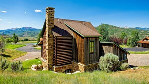 A rustic cabin with a stone chimney, wooden walls, and a green door, surrounded by a lush landscape and mountains in the background. - Great Cabin Experience - Stay Near to Aspen, AC (Woody Creek)