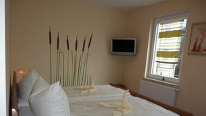 Un dormitorio con cama, almohadas, lámpara de noche, televisión y ventana con persianas.