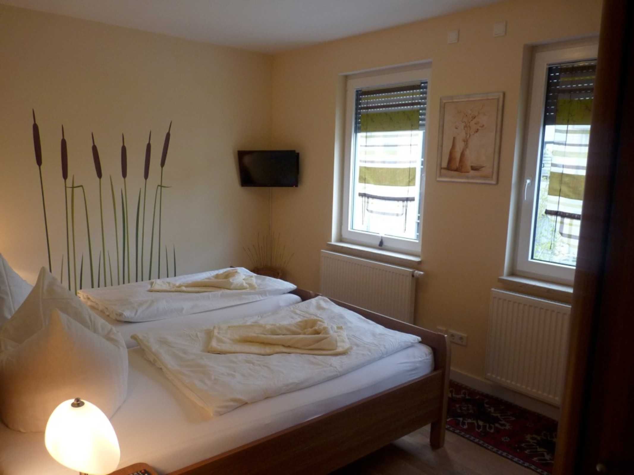 A bedroom with two beds, a TV mounted on the wall, and windows with blinds.