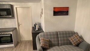 Ein gemütlicher Wohnraum mit Küchenzeile, einer Couch mit Karokissen, einer Kaffeemaschine und einem Sonnenuntergangsbild an der Wand.