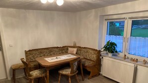 Ein Eckzimmer mit Holztisch, vier Stühlen, einem blumigen Sofa, einem Fenster mit Vorhängen und einer Heizung.