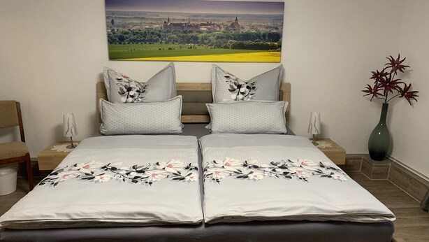 Ein Doppelbett mit floraler Bettwäsche, zwei Kissen, ein Nachttisch, eine Lampe, eine Vase mit roten Blumen und ein Landschaftsbild an der Wand.