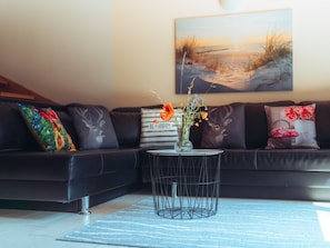 Un salon moderne avec un canapé en cuir noir, une table basse ronde ornée d’un vase de fleurs et un tableau représentant une scène de plage accroché au mur.