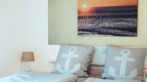 Une chambre à coucher avec un lit, des oreillers ornés de motifs d’ancres, une lampe de chevet et un tableau accroché au mur représentant un coucher de soleil sur l’océan.