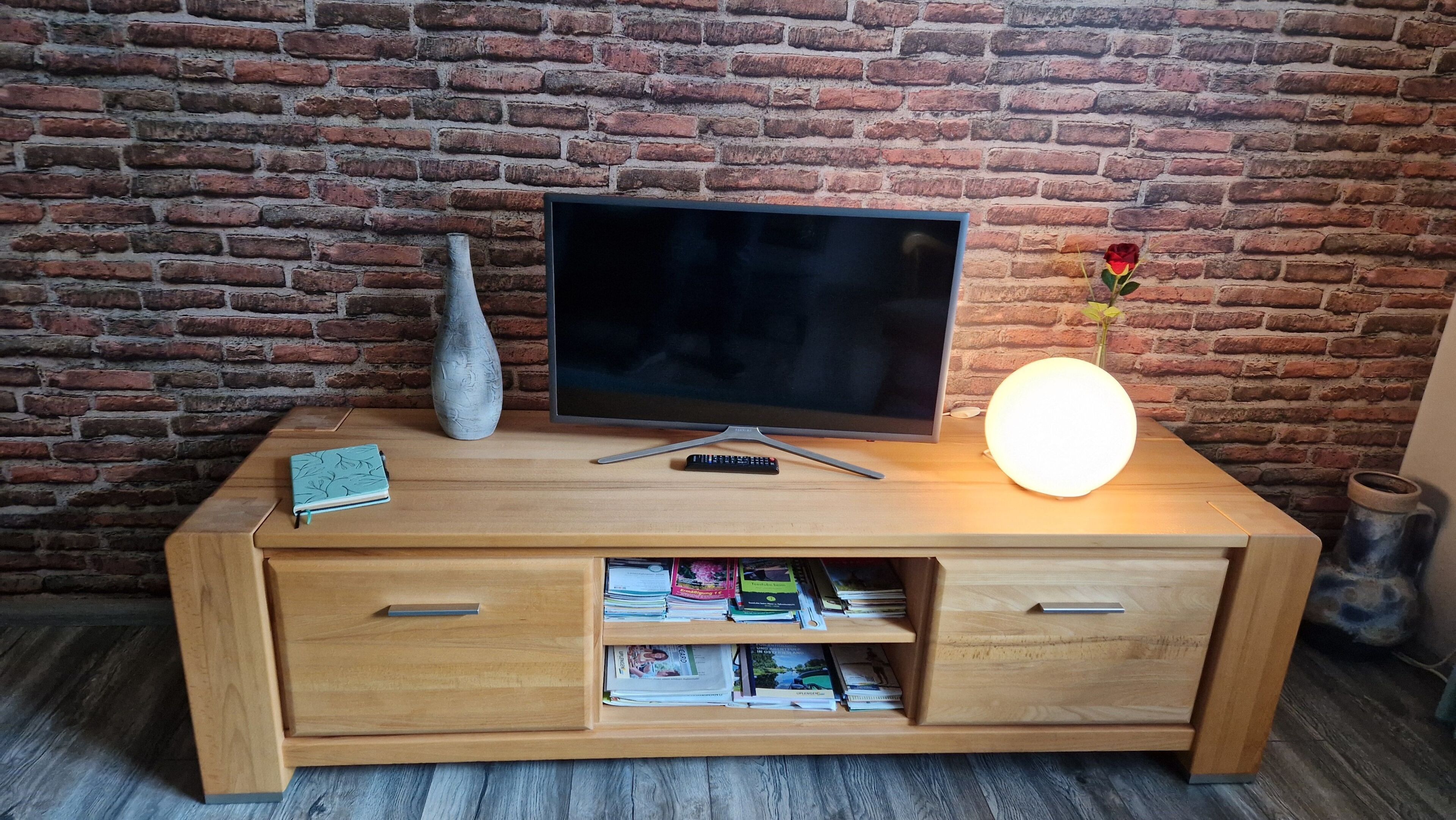 Un meuble de rangement en bois abritant un téléviseur à écran plat, un vase, une télécommande et une lampe allumée, le tout placé devant un mur en briques.