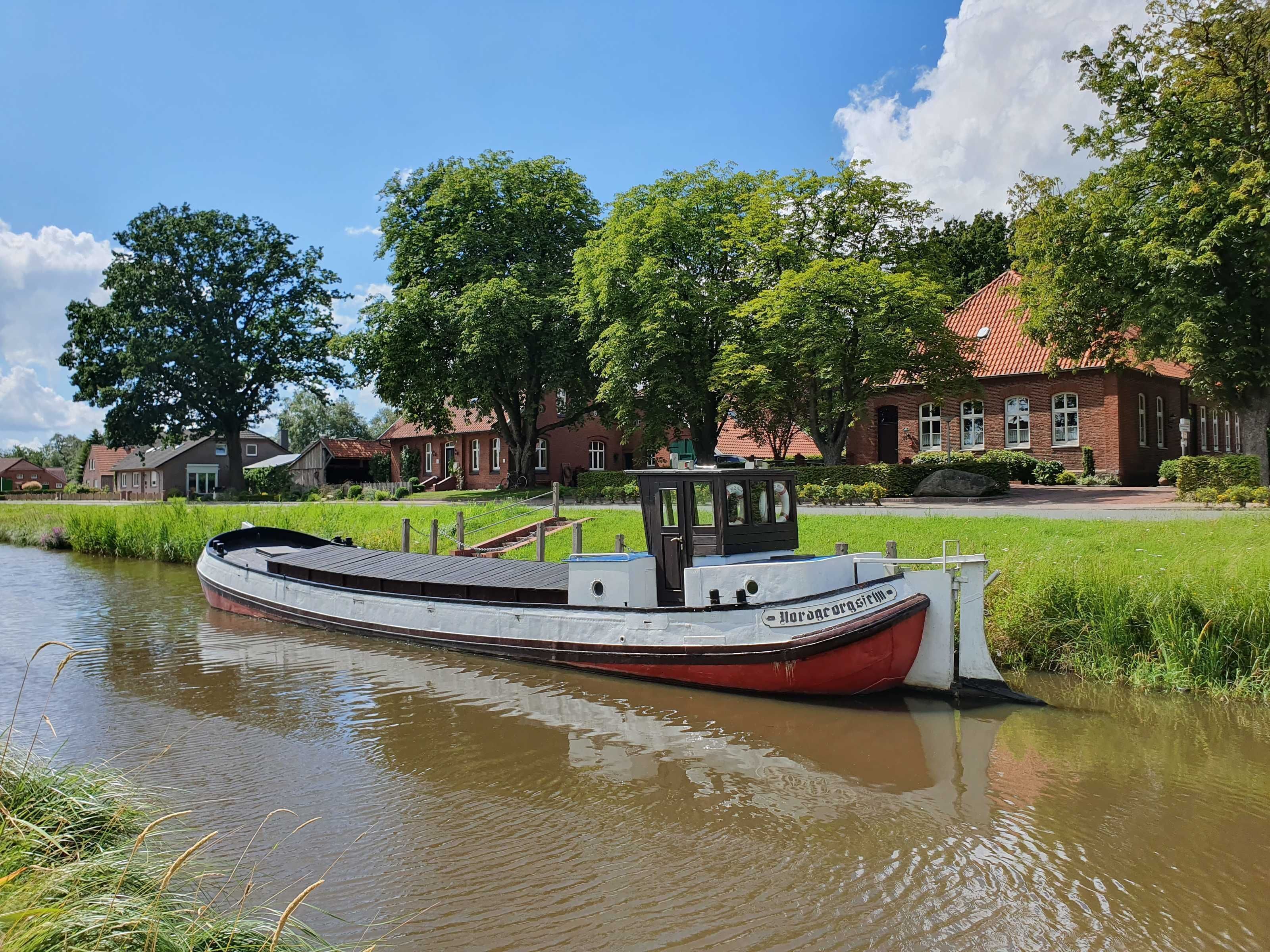 A narrowboat docked at a canal with a red and white structure, surrounded by greenery and traditional houses.