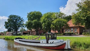 A narrowboat docked at a canal with a red and white structure, surrounded by greenery and traditional houses. - Ferienwohnung Kanalblick 35520 - Ferienwohnung Kanalblick (Uplengen)