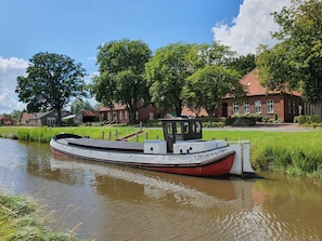 A narrowboat docked at a canal with a red and white structure, surrounded by greenery and traditional houses.