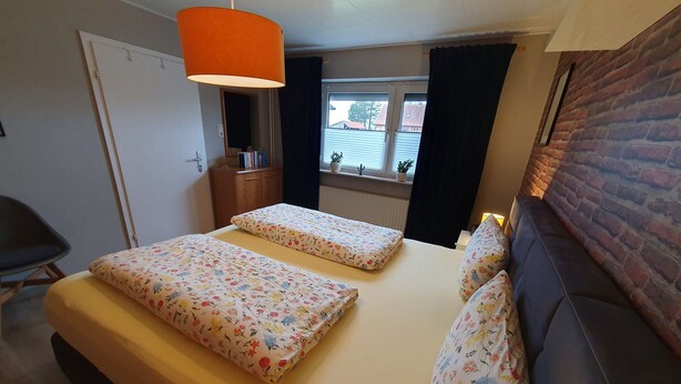 A bedroom with two beds, a sofa, a chair, a window with curtains, a brick wall, and a hanging lamp. - Ferienwohnung Kanalblick 35520 - Ferienwohnung Kanalblick (Uplengen)