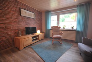 A living room with a brick wall, wooden entertainment unit, television, rocking chair, and a window with curtains.