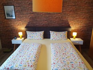 Una cama doble con ropa de cama floral, dos lámparas de noche, un reloj y un cuadro en una pared de ladrillo.