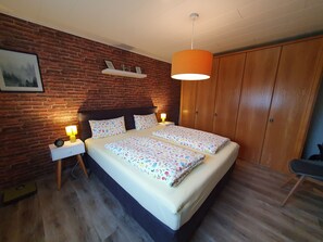 A double bed with floral bedding, a brick wall, a wooden wardrobe, and a hanging lamp.