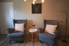 Two armchairs with pillows, a small round table with a vase, a wall-mounted deer head artwork, a floor lamp, and a door.