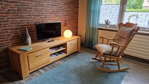 Ett vardagsrum med en tegelvägg, en träinredd TV-möbel, en gungstol och ett fönster med gardiner.