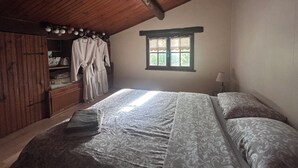 Een slaapkamer met een houten plafond, een bed met beddengoed met een patroon, een raam met gordijnen en een houten garderobe met daarin hangende gewaden.