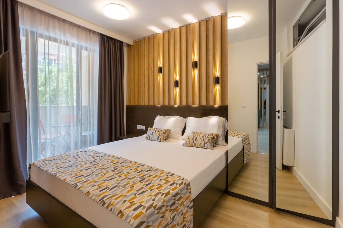A modern hotel room with a large bed, a wooden headboard, and a view of the outdoors through sheer curtains.