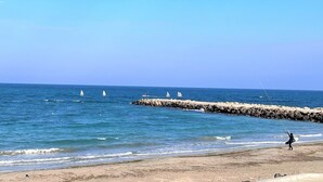 Una playa con un rompeolas rocoso y veleros a lo lejos.