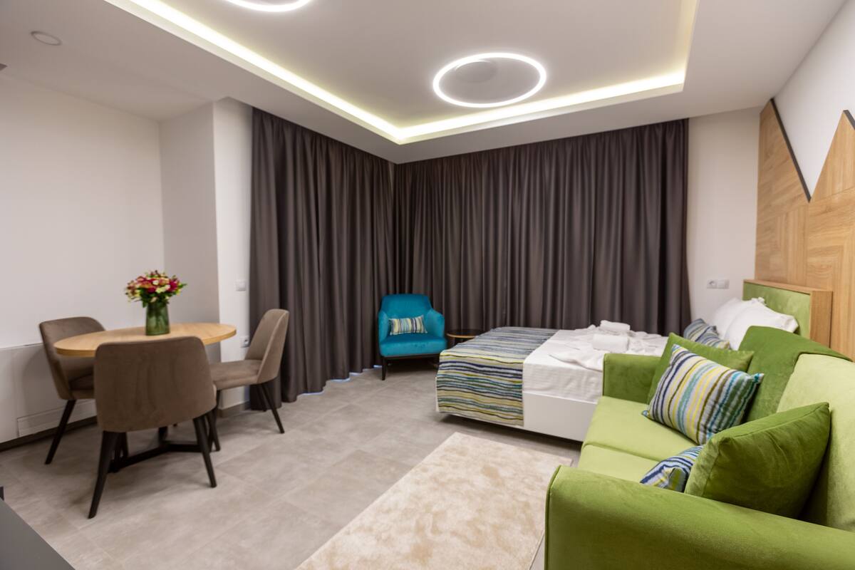 A modern hotel room with a dining area, a bed with green bedding, a blue armchair, and a wooden accent wall.