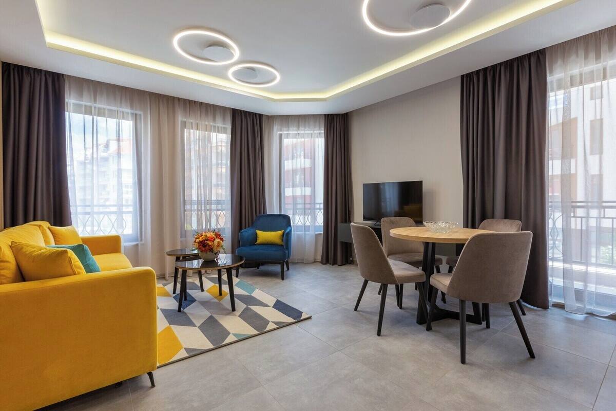 A modern living room with a yellow sofa, a blue armchair, a dining table with chairs, and a TV.