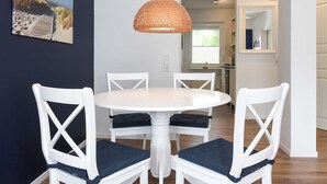 Un comedor con una mesa redonda blanca y sillas, una lámpara colgante tejida y un cuadro con una escena de playa en la pared.