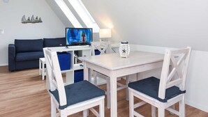 Ein modernes, helles Zimmer mit Esstisch, Stühlen, Fernseher und einer Couch.