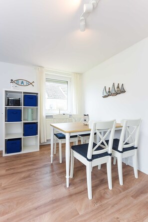 Um quarto com piso de madeira, paredes brancas, uma mesa branca com duas cadeiras, uma janela com cortinas e um aparador branco com caixas de armazenamento azuis.