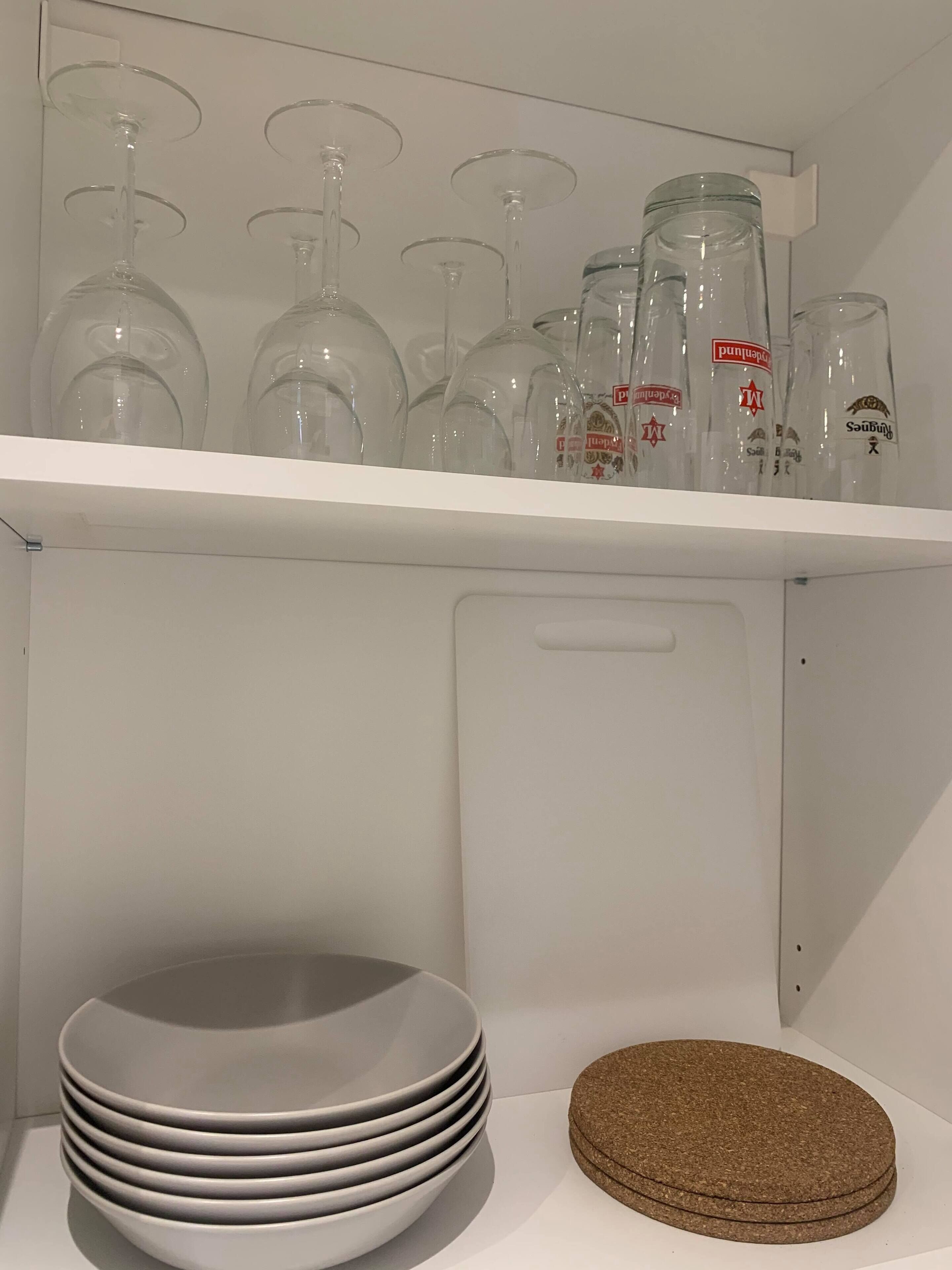 A kitchen shelf with wine glasses and bottles, a stack of white plates, and a cork coaster.