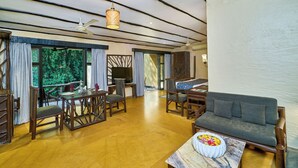 A spacious room with a wooden floor, a sofa, a dining table with chairs, a ceiling fan, and large windows offering a view of greenery.