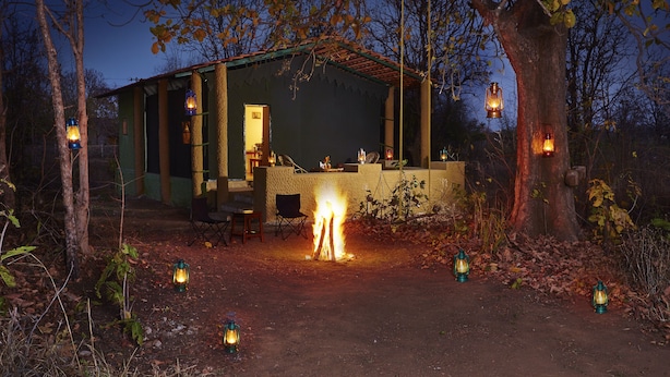 A rustic cabin with a fire in the center, surrounded by trees and multiple lanterns.
