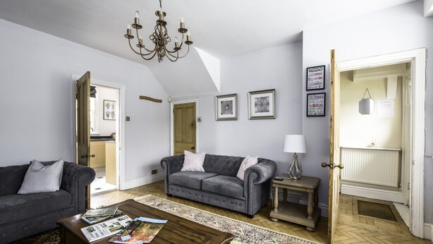 A living room with a grey sofa, a wooden coffee table, and a chandelier. There are framed pictures on the wall and a door leading to another room. - The cottage is a short walk to the Station, with trains to Liverpool / chester,  (Wirral, Merseyside)
