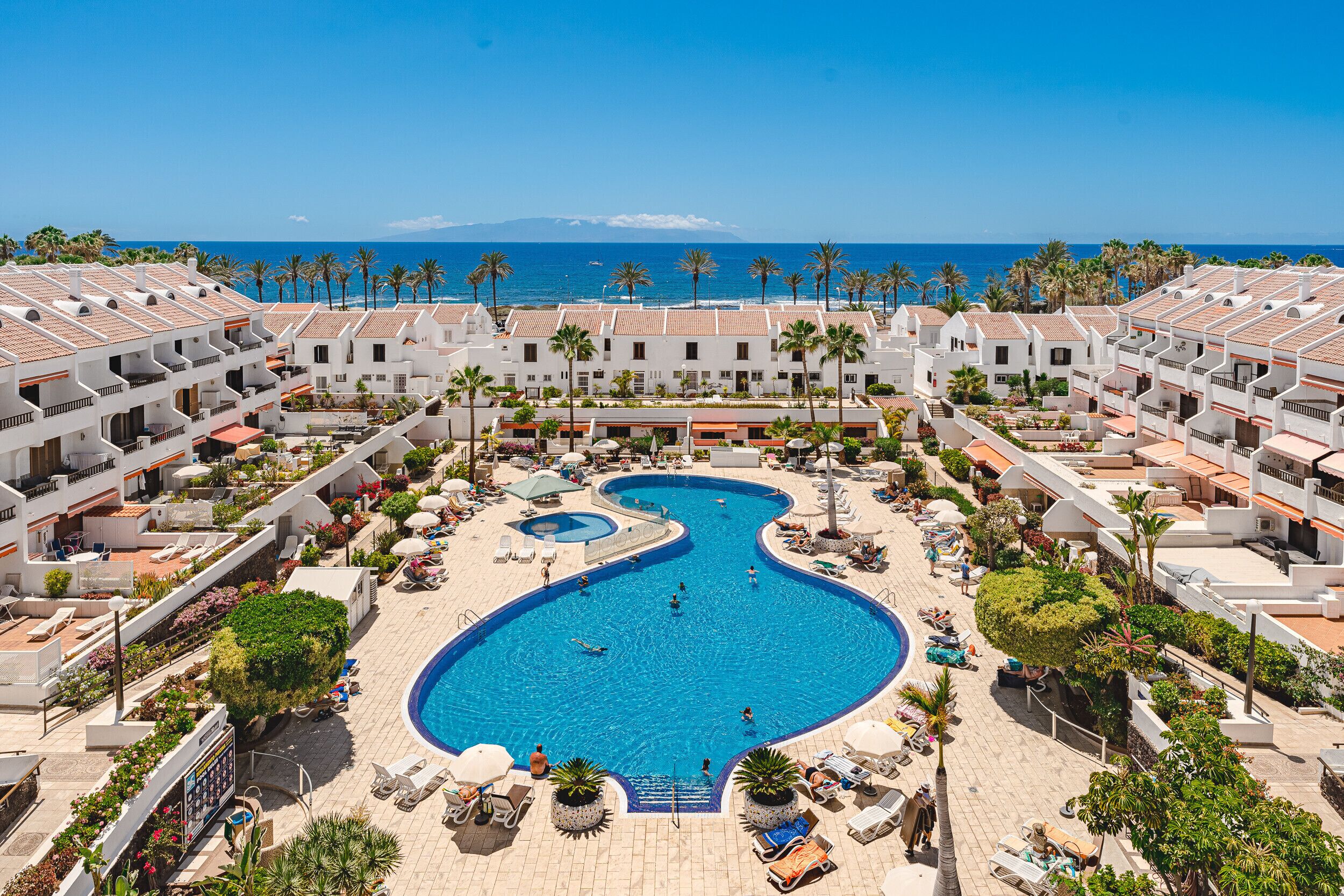 Un complexe hôtelier doté d’une piscine centrale, entourée de palmiers et de chaises longues, offrant une vue sur l’océan.