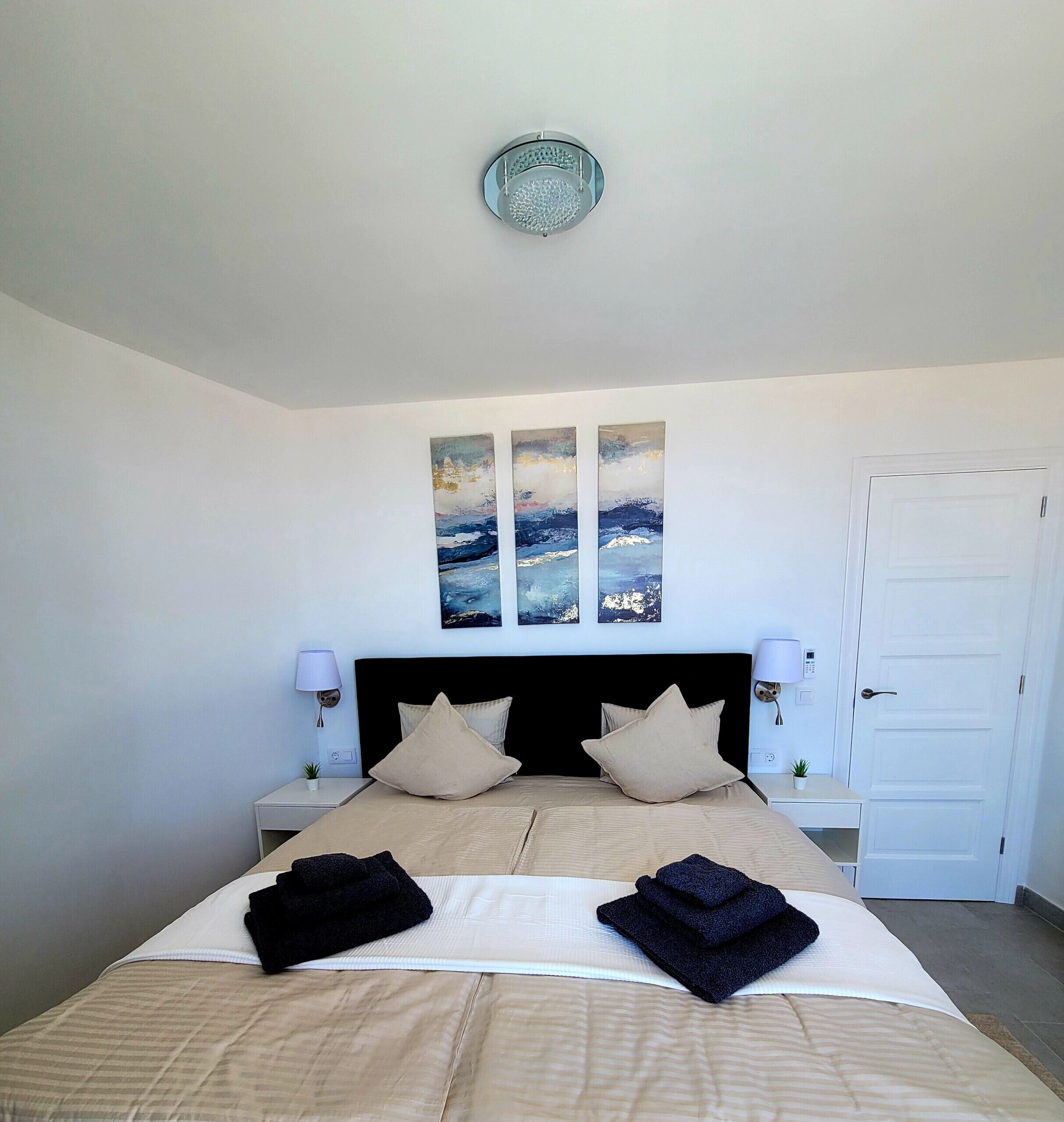 Quarto com cama, duas mesas de cabeceira, luminária na parede, uma porta e três quadros.