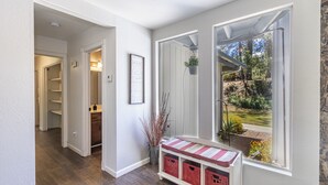 Um corredor com piso de madeira, um banco com cestos, uma moldura com uma citação inspiradora e vista para um jardim através de uma grande janela.