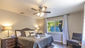 Quarto com cama, duas mesas de cabeceira com abajures, ventilador de teto, janela com cortinas e uma cadeira.